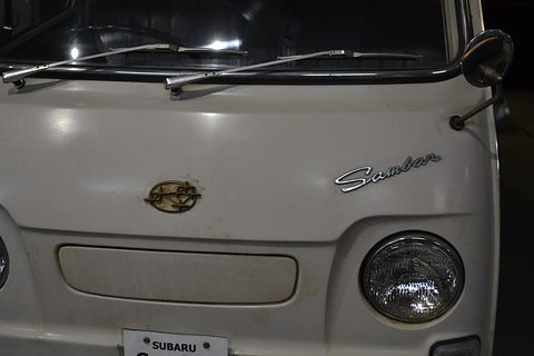 Close view of a cream van nose with a gold emblem, a chrome script badge and a round headlamp. Opens a larger view