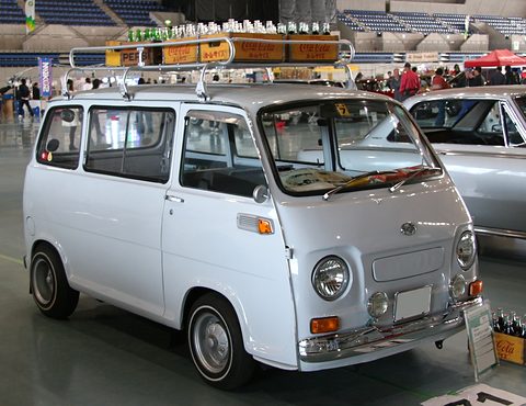 White small cab-over van with a roof rack loaded with crates and signs in an arena. Opens a larger view