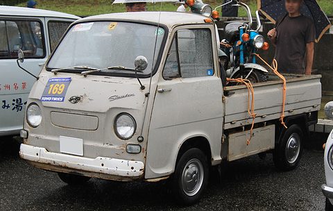 Weathered white small cab-over pick-up with a rally plate and a small motorcycle tied in the bed. Opens a larger view