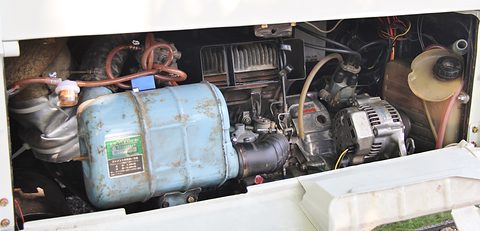 Open engine hatch showing a small engine, a blue air box, an alternator and hoses. Opens a larger view