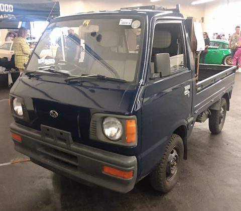 Navy small cab-over truck in a show hall, visitors and a green car behind. Opens a larger view