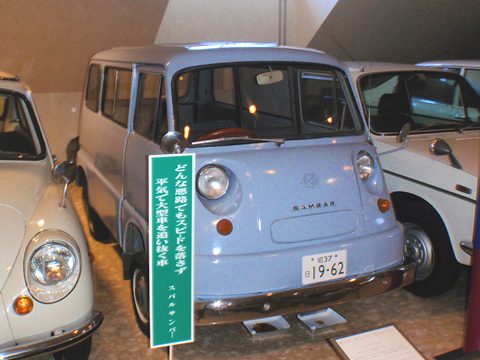 Pale blue small cab-over van with round headlamps behind a green museum sign, other small cars beside it.