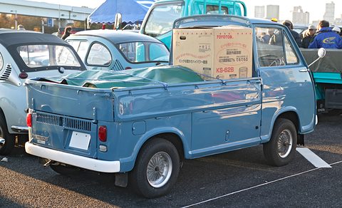 Rear of a blue small pick-up with a green load cover and a cardboard box, other small cars around. Opens a larger view