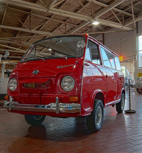 Red small cab-over van with chrome bumpers on a brick museum floor under steel roof trusses. Opens a larger view