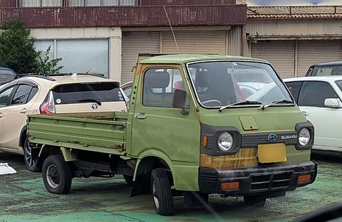 Faded green small cab-over truck with a dropside bed parked among modern cars. Opens a larger view