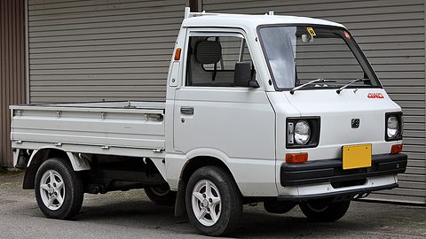 White small cab-over truck with a dropside bed and a yellow plate parked before a shutter. Opens a larger view