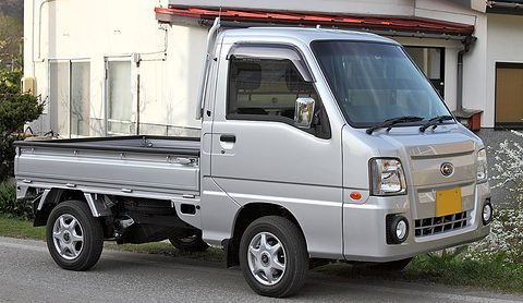 Side view of a silver small cab-over truck with a low dropside bed parked before a house. Opens a larger view