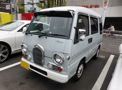 Pale blue retro-styled small van with round headlamps and a tall chrome grille in a car park. Opens a larger view