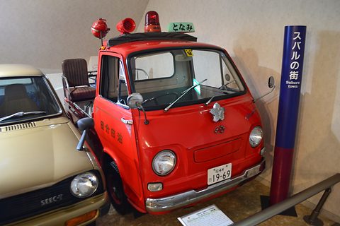 Red small cab-over truck with roof beacons and a ladder rack beside a blue museum pillar. Opens a larger view