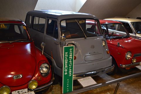 Grey small cab-over van with round headlamps beside a green sign, red small cars on either side. Opens a larger view
