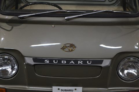 Close view of an olive-grey cab nose with a chrome SUBARU bar between two round headlamps. Opens a larger view