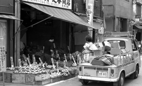 A small pick-up piled with boxes parked outside a fruit shop, a woman standing in its bed.