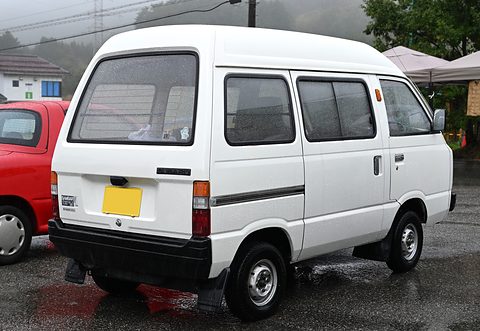 Rear of a white high-roof small van with a yellow plate on wet tarmac. Opens a larger view