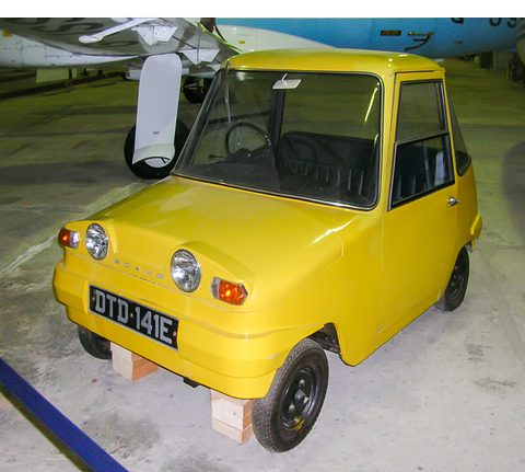 Yellow tiny electric car on wooden blocks, an aircraft nose above and behind it.
