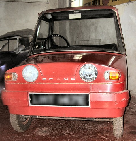 Faded red tiny electric car head-on in a whitewashed shed, an old car beside it.