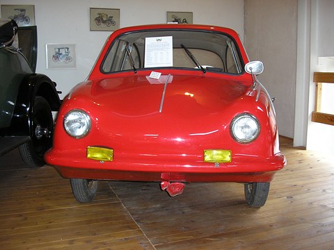 Red rounded small car with yellow fog lamps on a wooden floor, a display card on its windscreen. Opens a larger view