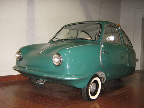 Dark green rounded three-wheeler with whitewall tyres and a chrome mirror on a brick-red floor.