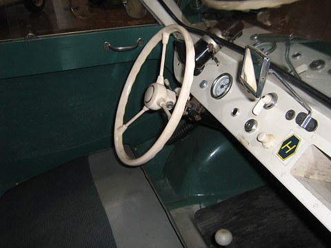 Cream two-spoke steering wheel before a white dashboard with one dial, toggles and an H badge, green trim. Opens a larger view