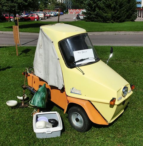 Yellow three-wheeler with a white canvas cover on grass, a price note on its windscreen, crockery beside it. Opens a larger view