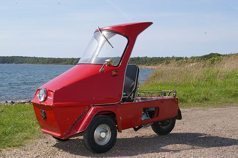 Red single-seat three-wheeler with a tall fibreglass canopy on gravel by a lake shore. Opens a larger view