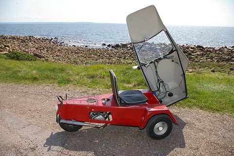 Red three-wheeler with its whole canopy tipped forward, showing a single seat and rear rack, by the sea.