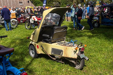 Rusty pale yellow three-wheeler with the canopy up, a rear rack and exhaust, crowds and motorcycles behind. Opens a larger view
