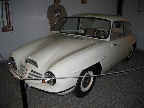 White rounded two-door coupé behind a rope barrier on a grey floor, a visitor standing behind.