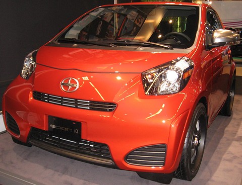 Orange city car with a black Scion grille and dark wheels on a show floor, seen nearly head-on. Opens a larger view
