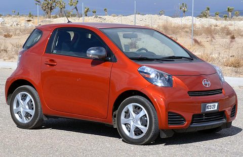 Orange city car with silver wheel covers parked on concrete, desert scrub and Joshua trees behind. Opens a larger view