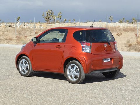 Rear of an orange city car with tall tail lamps on a concrete apron, desert and Joshua trees behind. Opens a larger view