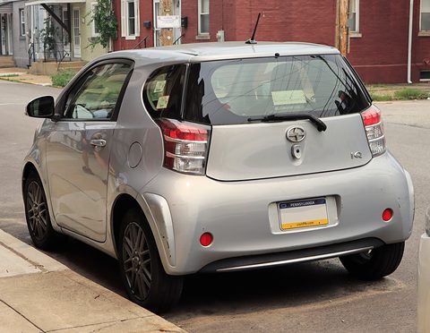 Rear of a silver city car with tall tail lamps parked on a street lined with brick houses. Opens a larger view