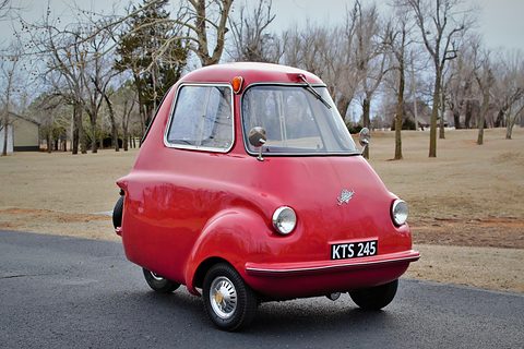 Tall narrow red three-wheeler on a tarmac drive, seen from the front left, bare trees behind. Opens a larger view