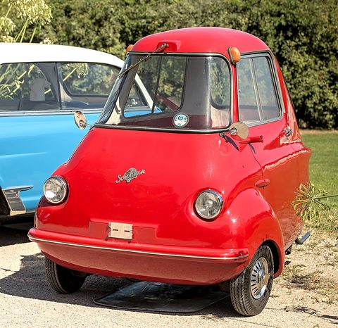 Red three-wheeler on gravel seen from the front, a blue car at left. Opens a larger view