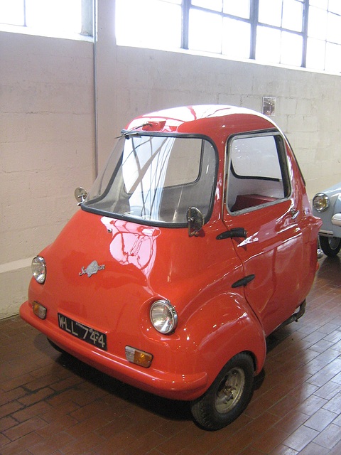 Red three-wheeler on a brick museum floor by a white wall, seen from the front left. Opens a larger view