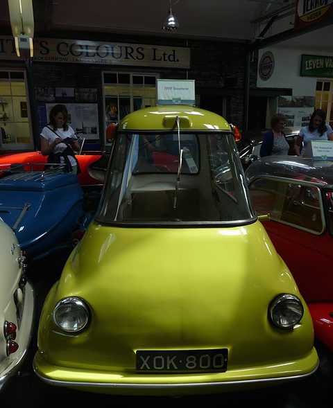 Lime green three-wheeler seen head-on in a museum, visitors and a shop sign behind. Opens a larger view