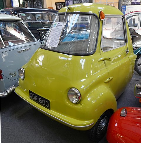 Lime green three-wheeler among other small cars in a museum, an information card on its windscreen. Opens a larger view