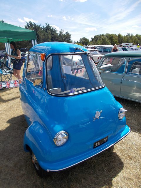 Bright blue three-wheeler on dry grass, a marquee and visitors behind. Opens a larger view