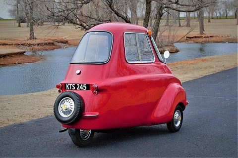 Rear of a tall red three-wheeler on a drive by a pond, spare wheel on the tail. Opens a larger view