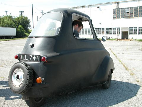 A tall narrow black microcar seen from behind, a spare wheel on its tail and a man looking out of its side window. Opens a larger view