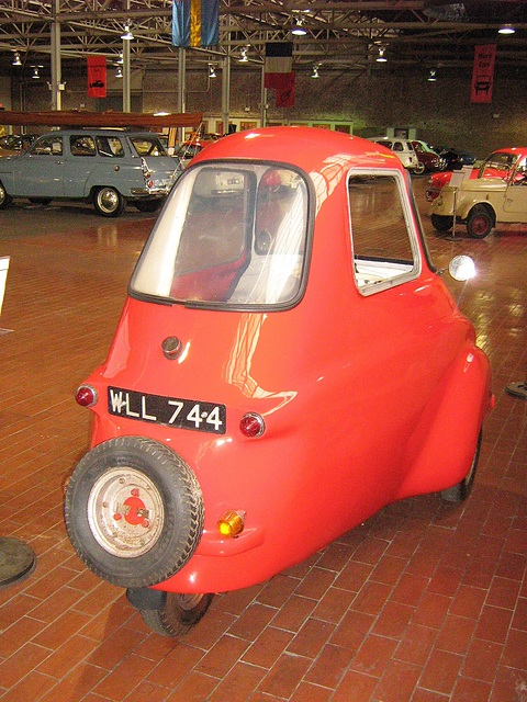 Red three-wheeler from behind with its spare wheel, other cars in the hall behind. Opens a larger view