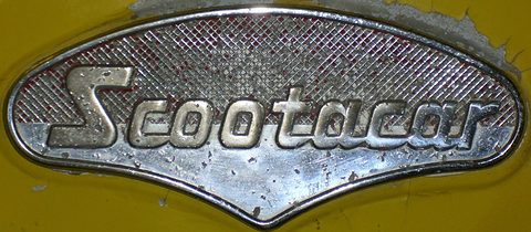 Chrome badge with the Scootacar script on a red-backed mesh, on yellow paint. Opens a larger view