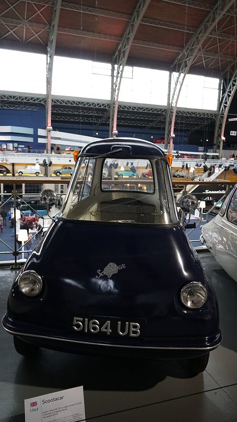 Dark navy three-wheeler seen from the front in a large hall with an iron roof. Opens a larger view