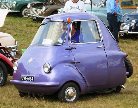 Purple three-wheeler in side view on grass with a man seated inside, people behind. Opens a larger view