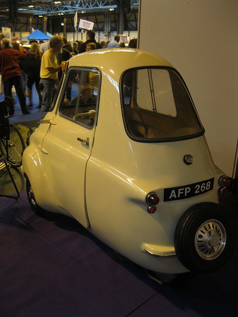 Rear three-quarter of a cream three-wheeler with a spare wheel on its tail, visitors behind. Opens a larger view