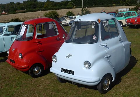 Red and pale blue three-wheelers side by side on grass, other classics behind