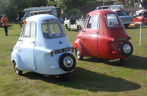 Two three-wheelers on grass seen from behind, a pale blue one and a red one. Opens a larger view