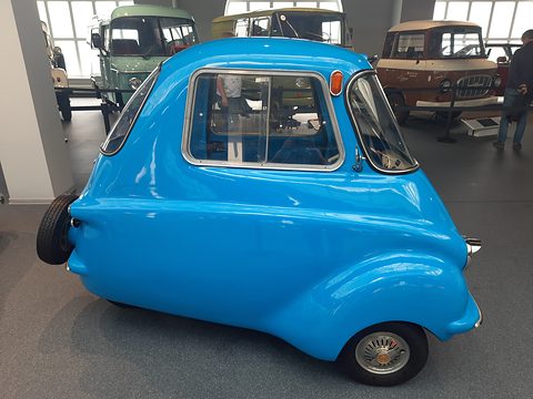 Bright blue three-wheeler in side view on a grey museum floor, vans behind
