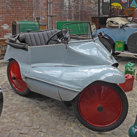 Side view of a grey three-wheeler with a black seat and red disc wheels on a cobbled floor.