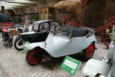 Side view of a grey three-wheeler with red wheels and a long tail, a museum label on a stand beside it. Opens a larger view