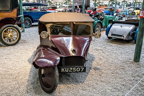 Front view of a maroon three-wheeler with a hooded cabin, two lamps and a single front wheel offset to one side.